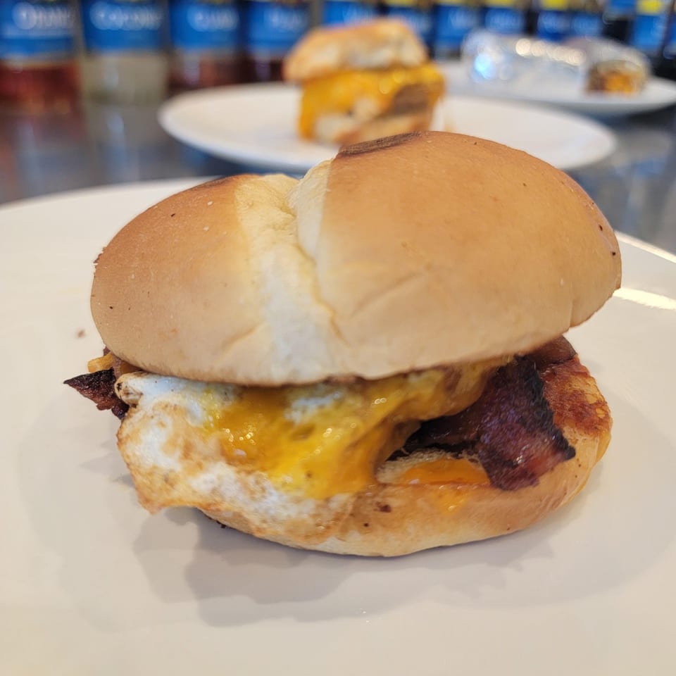 Close-up of a breakfast sandwich with egg, bacon, and melted cheese on a bun.
