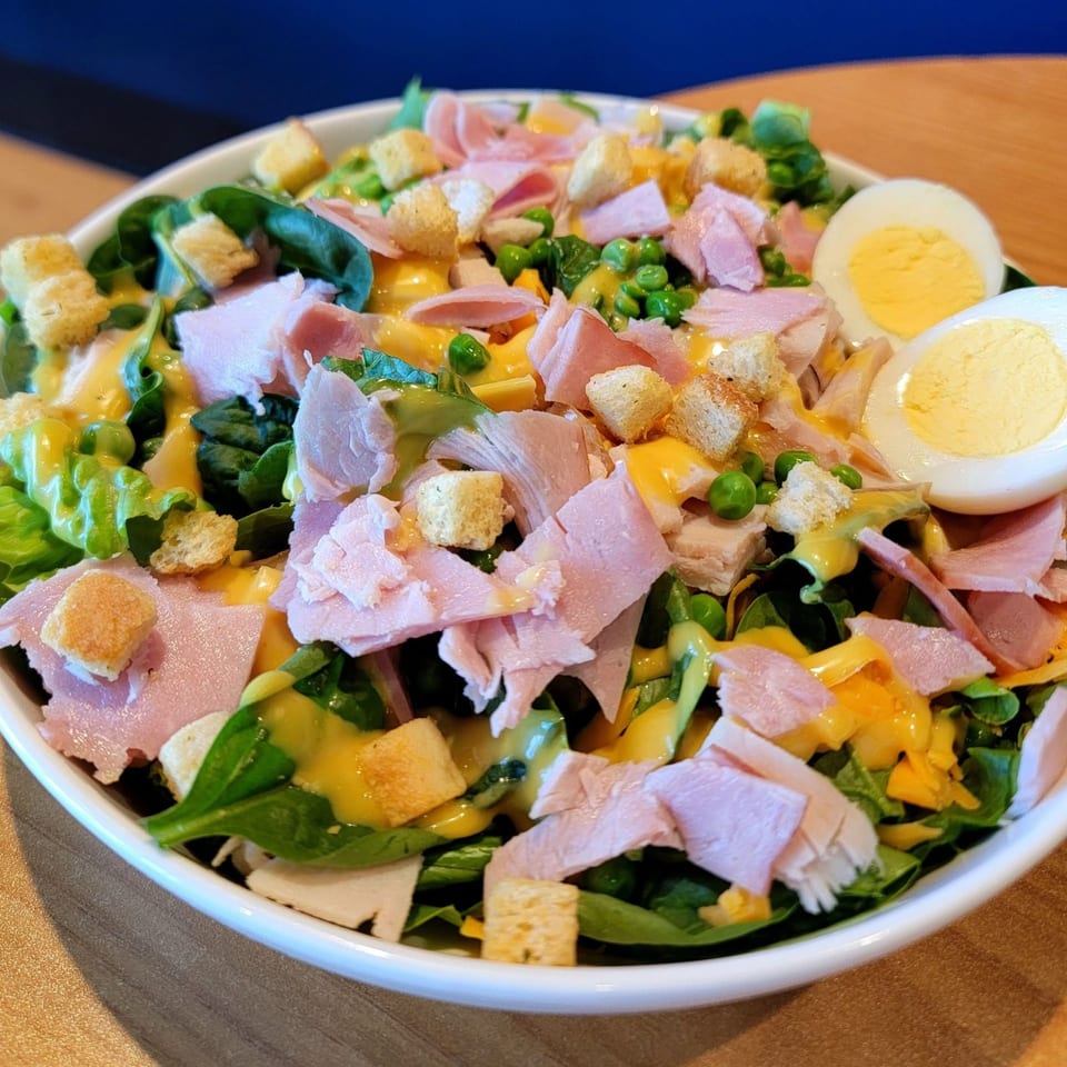 A vibrant chef salad with ham, hard-boiled eggs, croutons, peas, and dressing in a white bowl.